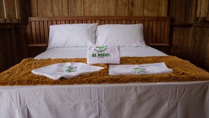 Una cama con sábanas blancas, dos almohadas y tres toallas con el logotipo 'EL NIDO' en un edredón marrón y mullido.