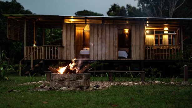 Una cabaña de madera con un brasero al frente, una terraza y un dormitorio con cama y mesita de noche.