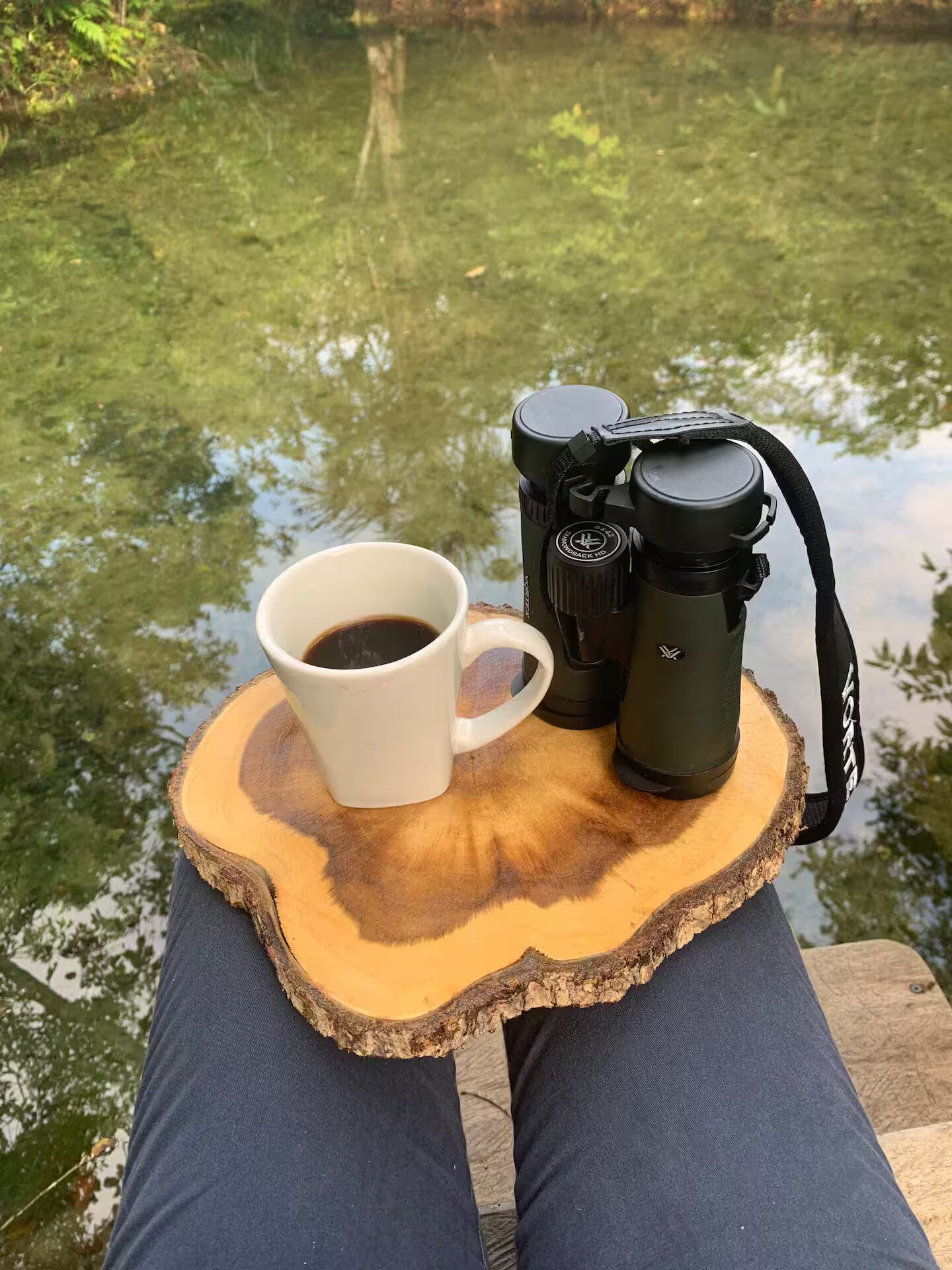 Une tasse blanche remplie de café, un appareil photo et un trépied, posés sur une surface en bois, près d’une étendue d’eau.
