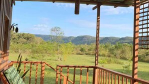 A wooden deck with a metal roof, a green cushioned chair, and a view of a green landscape with hills. - El Nido serene cabin with natural pool and chef #1 (La Macarena)
