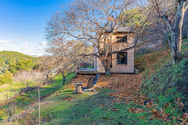 A wooden cabin with a deck, surrounded by trees and a hillside.