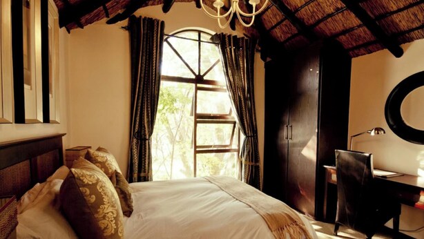 A bedroom with a bed, a desk, a chair, a window with curtains, and a chandelier. - Luxury 3 Apartment (Sandton)