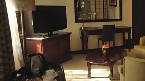 Ein Hotelzimmer mit Flachbildfernseher auf einer Holzkommode, einem Esstisch mit Stühlen, einem kleinen runden Tisch mit Lampe und einem Sofa.