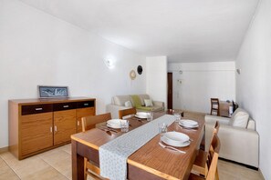 Un coin repas avec une table en bois dressée pour quatre personnes, un canapé, un buffet et, au fond, une petite cuisine.