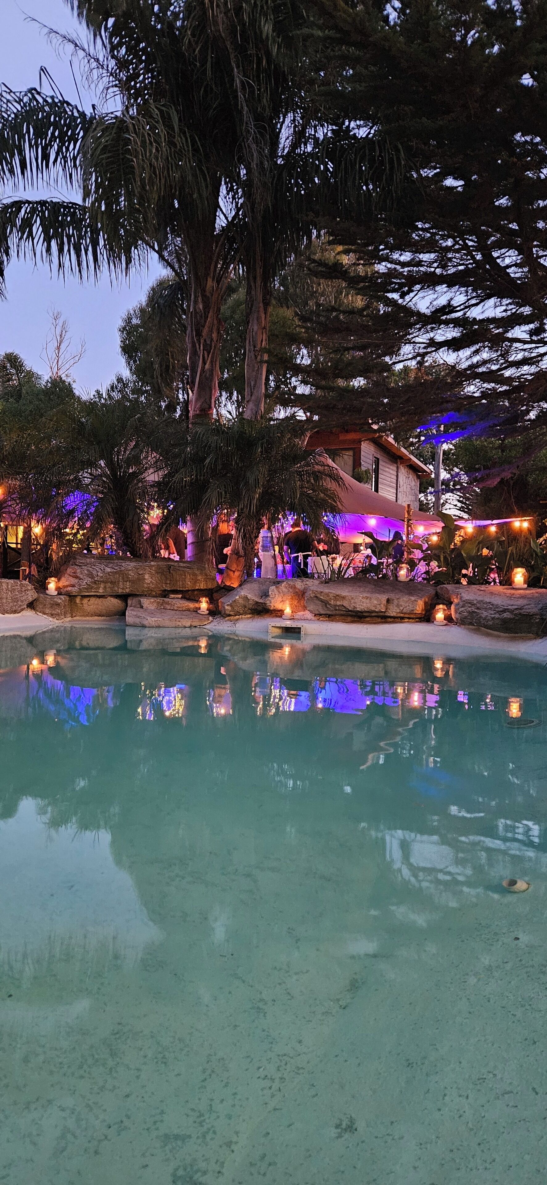 Uma área de piscina com água cristalina, rodeada por árvores e um edifício com iluminação externa.
