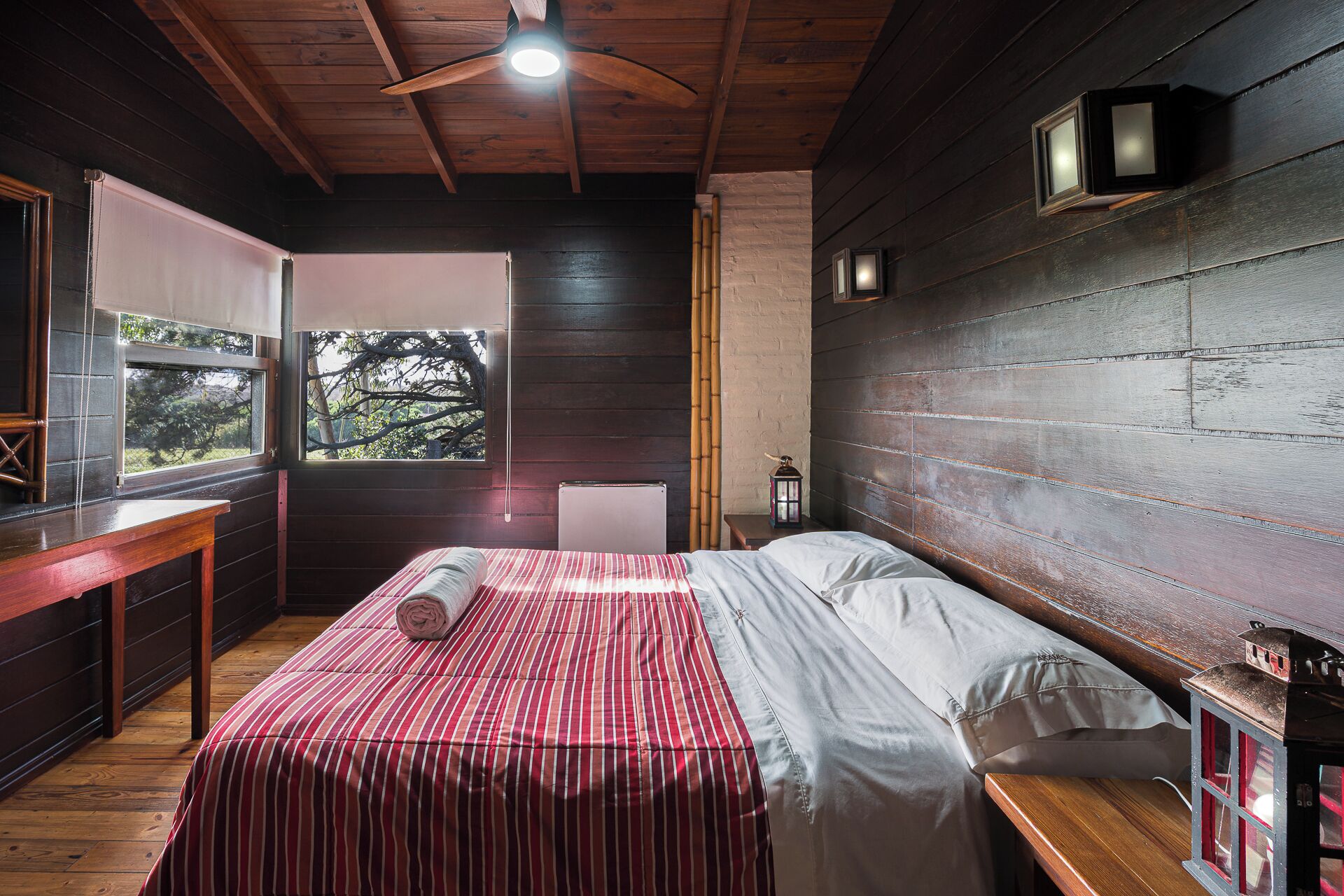 Una habitación con una cama, una ventana, techo de madera y una lámpara de pared.