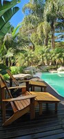 Un terrazzo in legno con poltrone relax affacciato su una piscina circondata da vegetazione tropicale.