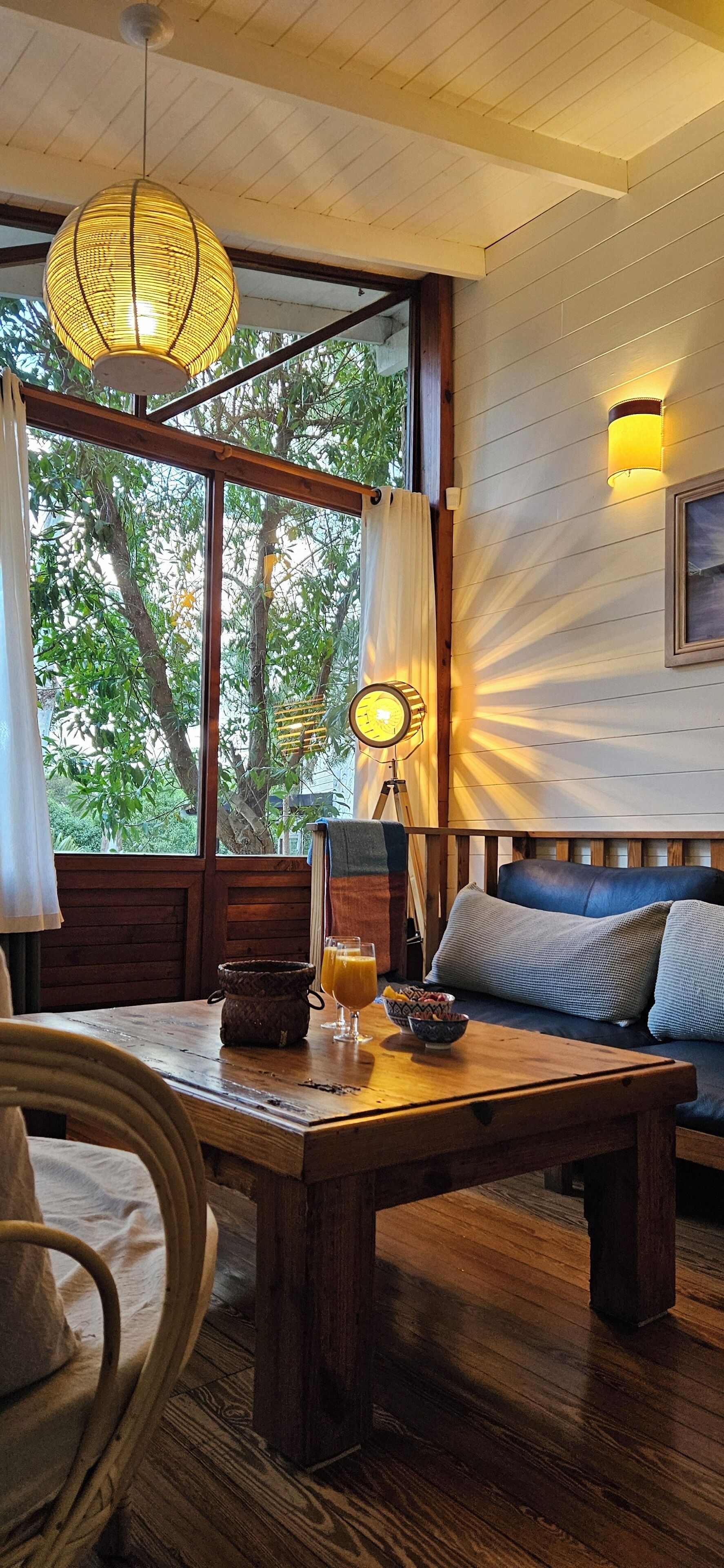 Una habitación acogedora con una mesa de madera, un sofá y un amplio ventanal con vistas a árboles.