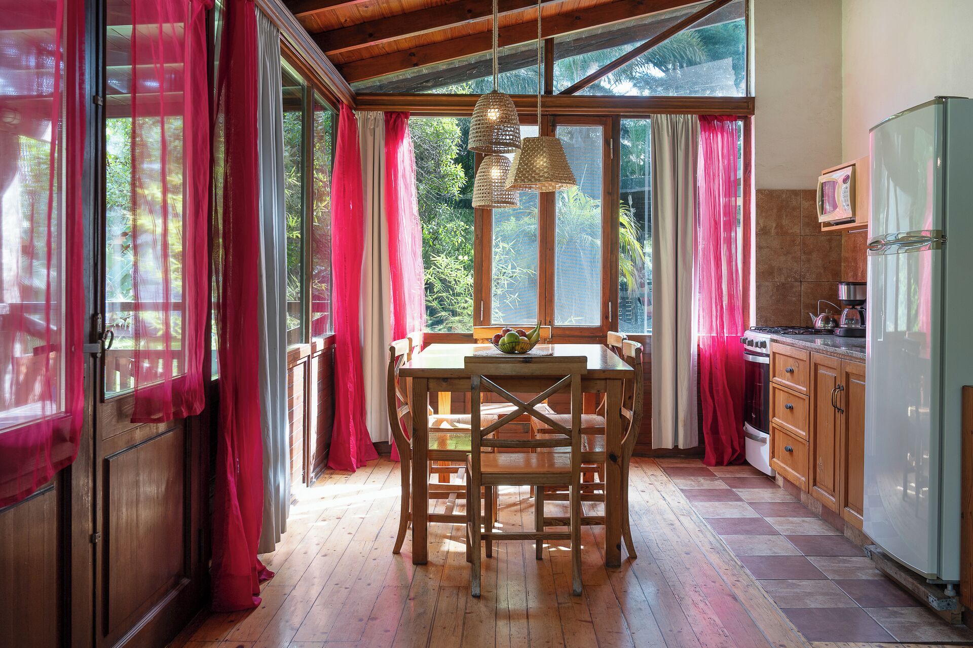 A kitchen with wooden cabinets, a dining table with chairs, a refrigerator, and a window with curtains.