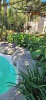 A pool surrounded by rocks and lush greenery, with a wooden deck and a stone building in the background.