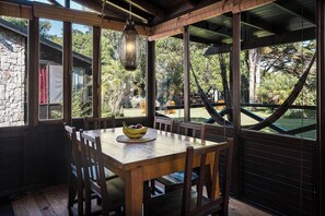 A wooden dining table with chairs, a hanging light fixture, and a view of a garden through large windows. - Complejo Aramis Chapadmalal (Colonia Chapadmalal)