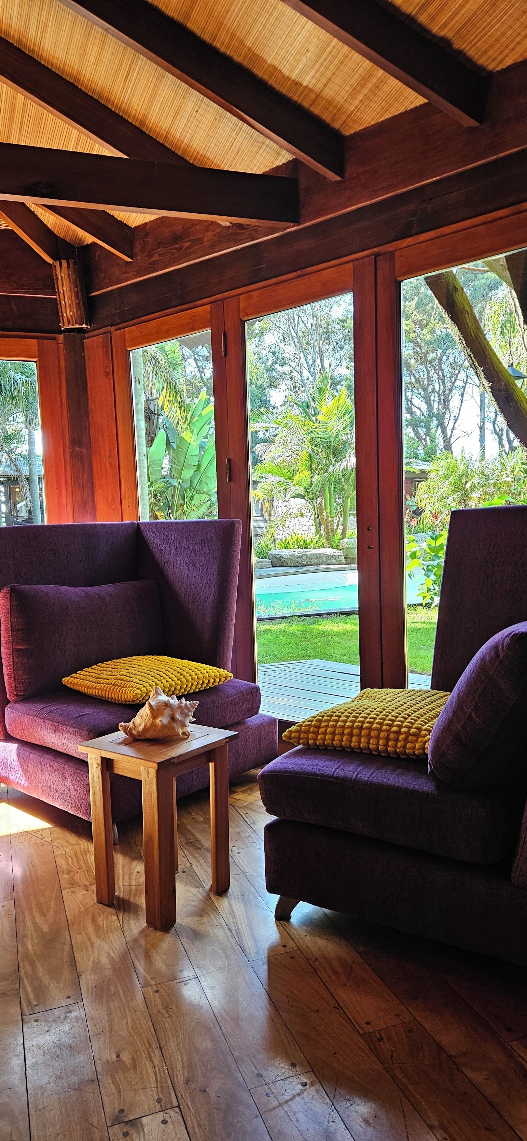 A room with two purple sofas, a wooden coffee table, and large windows with a view of greenery.