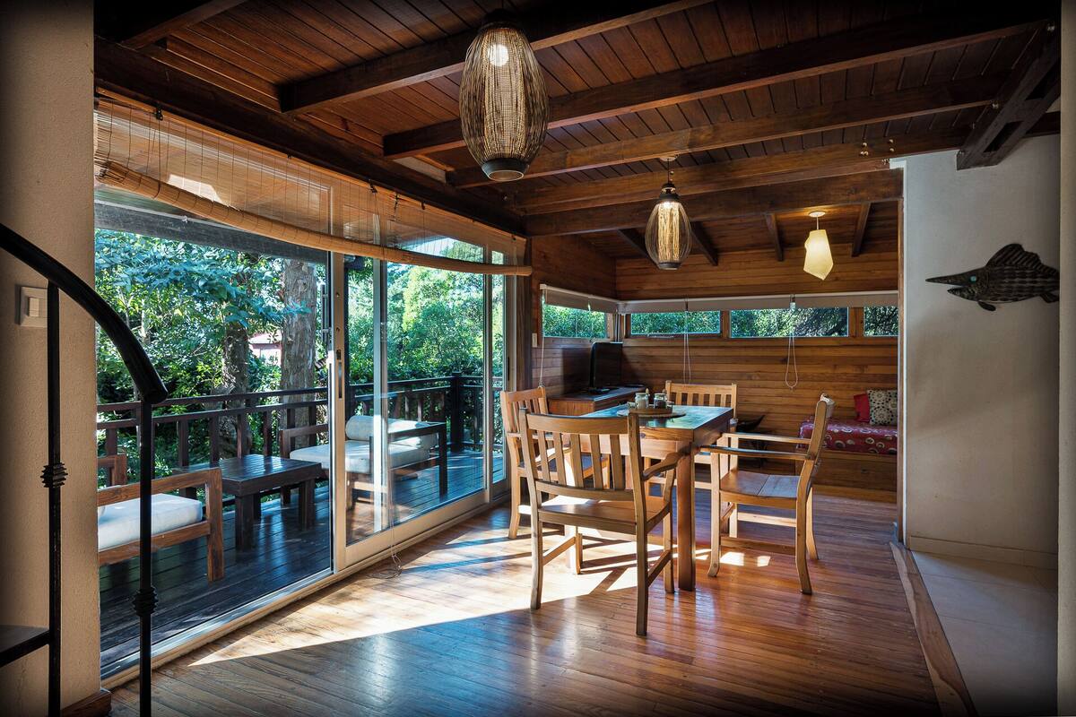 A spacious room with a wooden floor, a dining table with chairs, a large window, and a ceiling with wooden beams and hanging lights.