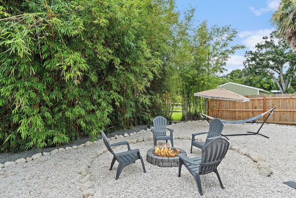 An outdoor seating area with a fire pit and chairs, surrounded by a gravel patio and dense foliage.
