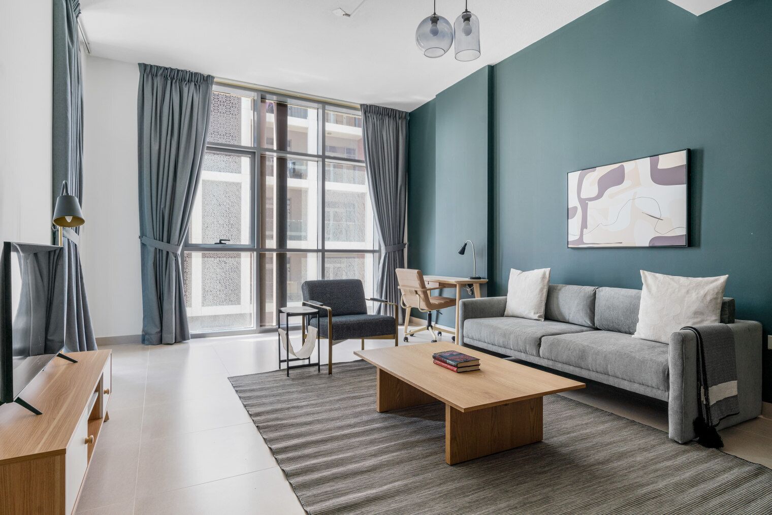 A modern living room with a grey sofa, a wooden coffee table, a desk with a chair, and large windows with grey curtains.