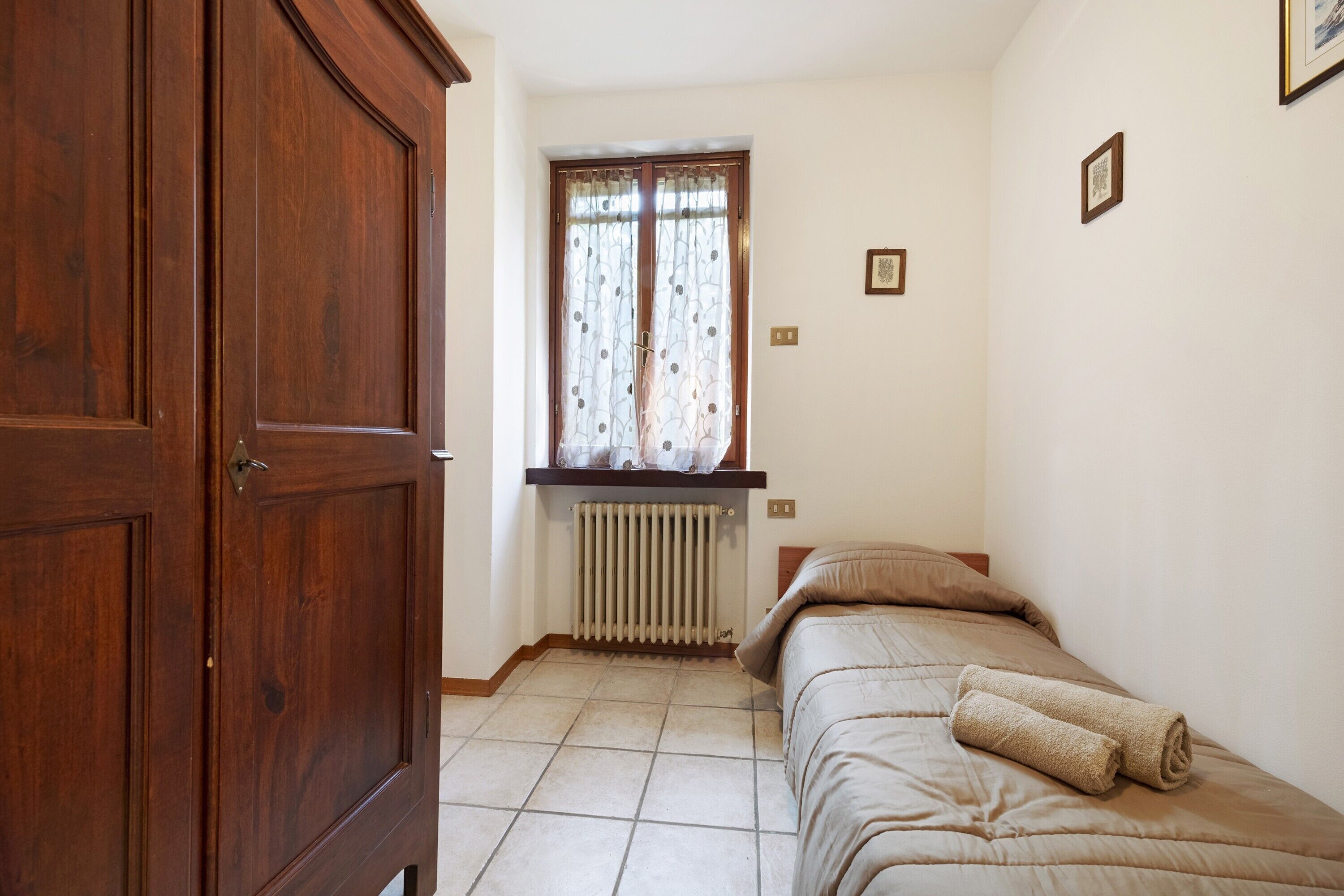Une chambre avec une armoire en bois, un radiateur, une fenêtre avec des rideaux, un lit avec une serviette roulée et un tableau encadré au mur.