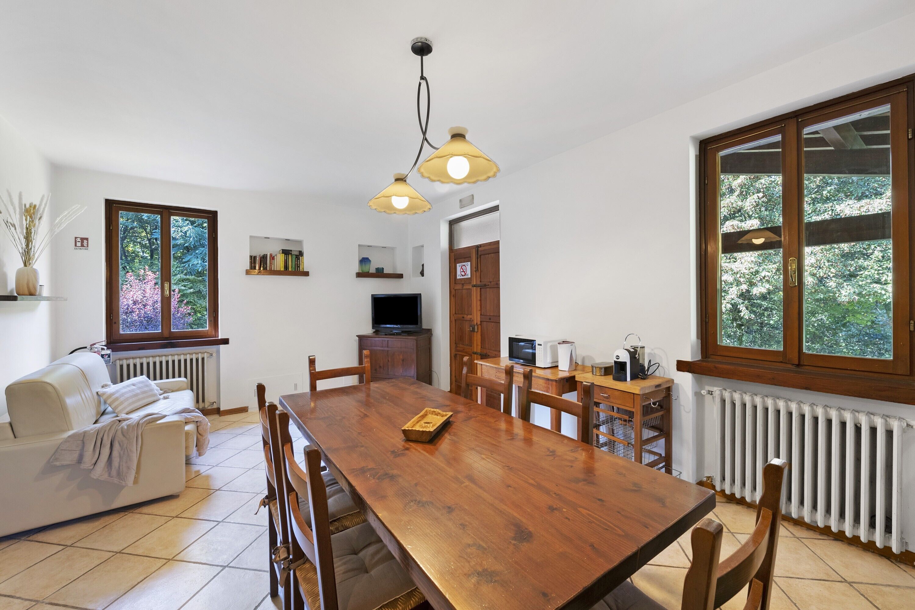 Un coin repas comprenant une table en bois, des chaises, un canapé, un téléviseur et une fenêtre donnant sur des arbres.