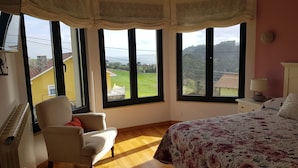A bedroom with a large bed, a chair, and a view of a green landscape through three windows.