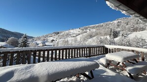A snow-covered deck with wooden railings and benches, overlooking a snowy landscape with houses and trees.