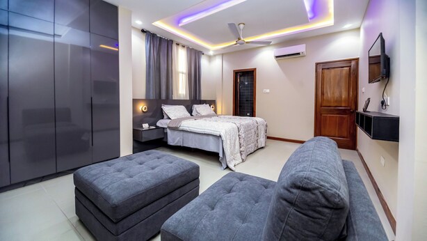 Une chambre à coucher moderne avec un lit, des poufs gris, un canapé, un ventilateur de plafond et un téléviseur.