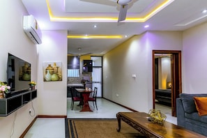 A modern living room with a flat-screen TV, a dining area with chairs and a table, and a sofa with a cushion.