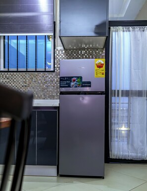 Une cuisine équipée d’un réfrigérateur, d’une fenêtre avec des stores, d’un crédence carrelée et d’un rideau.