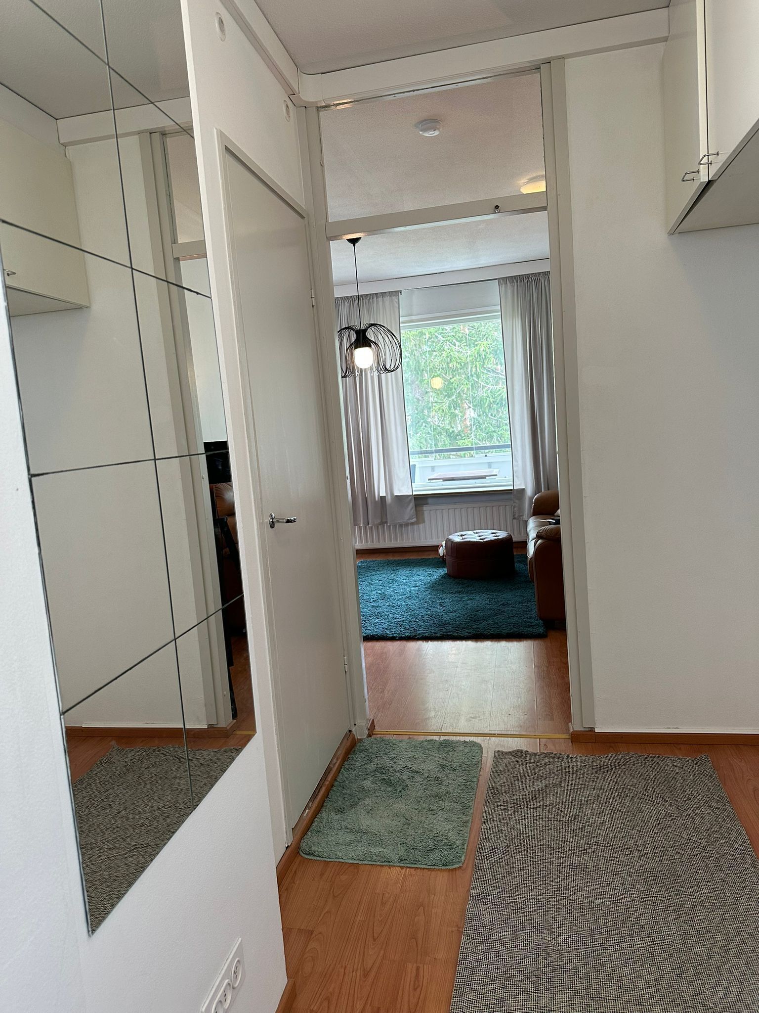 A hallway with a wooden floor, a green rug, a patterned rug, a window with curtains, and a ceiling light fixture.