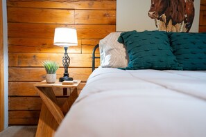 A wooden bed with white bedding and teal pillows, a bedside lamp, and a small potted plant on a wooden nightstand.