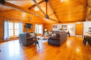 A spacious log cabin interior with a high ceiling, wooden floors, a fireplace, a television, and a dining area.