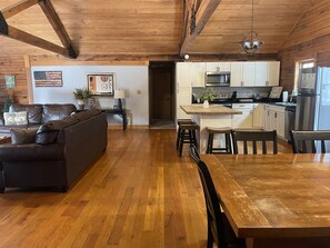 A spacious living area with a wooden floor, a dark brown sofa, and a dining table with chairs. A kitchen with white cabinets and stainless steel appliances is visible in the background.