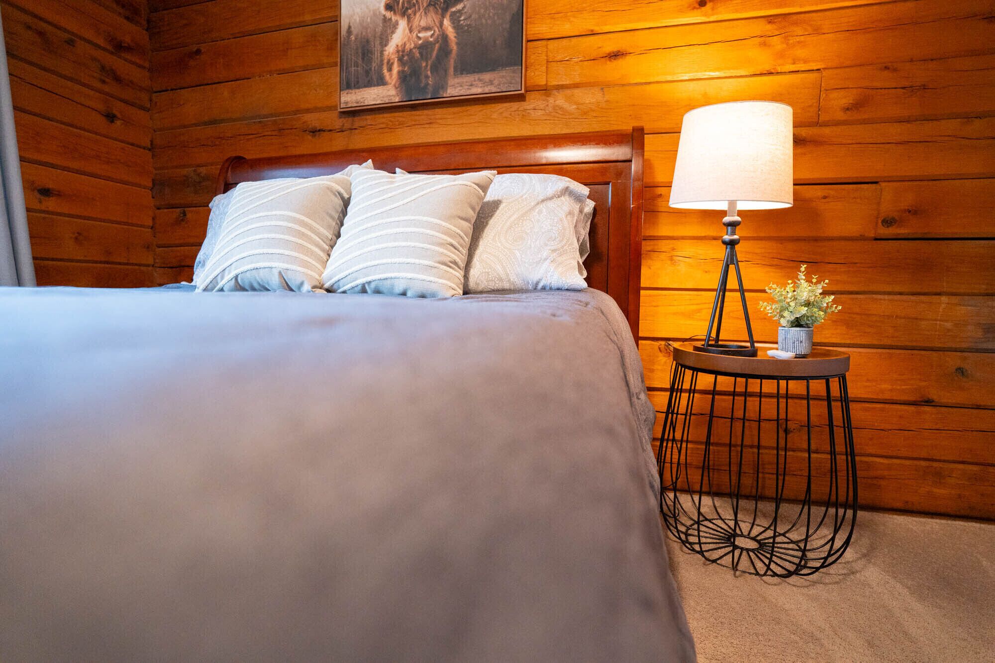 Una habitación tipo cabaña de madera con una cama, una lámpara y una pequeña mesa con una planta.