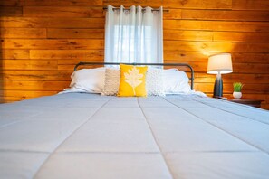 Ein Zimmer mit Holzwänden, einem Bett, einem Nachttisch mit Lampe und einem mit Vorhängen drapierten Fenster.