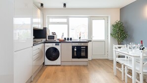 A modern kitchen with white cabinetry, a built-in washing machine, an oven, and a dining area with a white table and chairs. - Open Mind Property - 8 Guests, 4 Beds & Free Parking (Hayling Island)