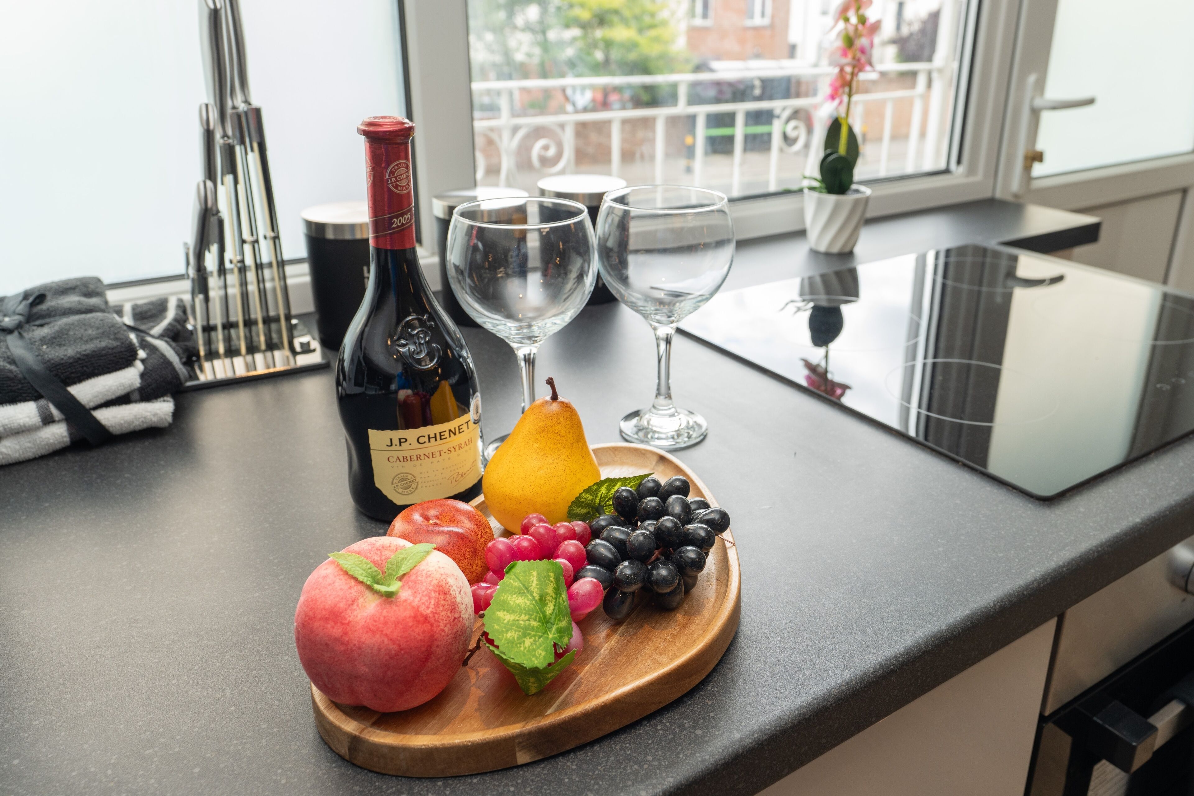 Un comptoir de cuisine avec une bouteille de vin, deux verres, une poire, des raisins et une planche en bois sur laquelle sont disposés des fruits.