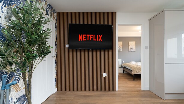 A modern room with a wooden panel wall, a mounted television displaying "NETFLIX," and a bed in the background. - Open Mind Property - 8 Guests, 4 Beds & Free Parking (Hayling Island)