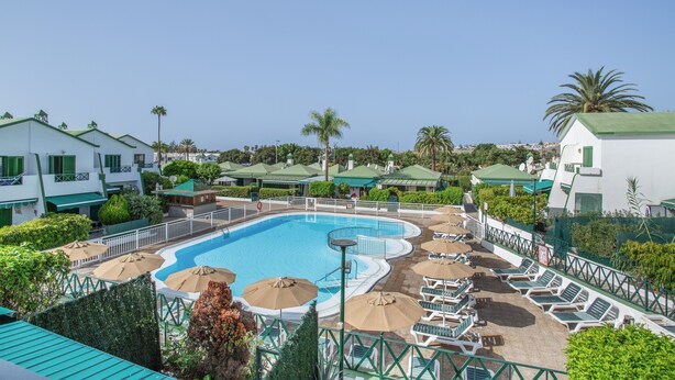 A swimming pool with sun loungers and umbrellas, surrounded by greenery and white buildings with green roofs.
