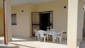 A covered outdoor area with a table and chairs, a door with a metal gate, and a small flower vase.