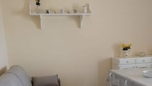 A neutral-toned living room with a grey sofa, a dining table with chairs, and a white shelf with decorative items.