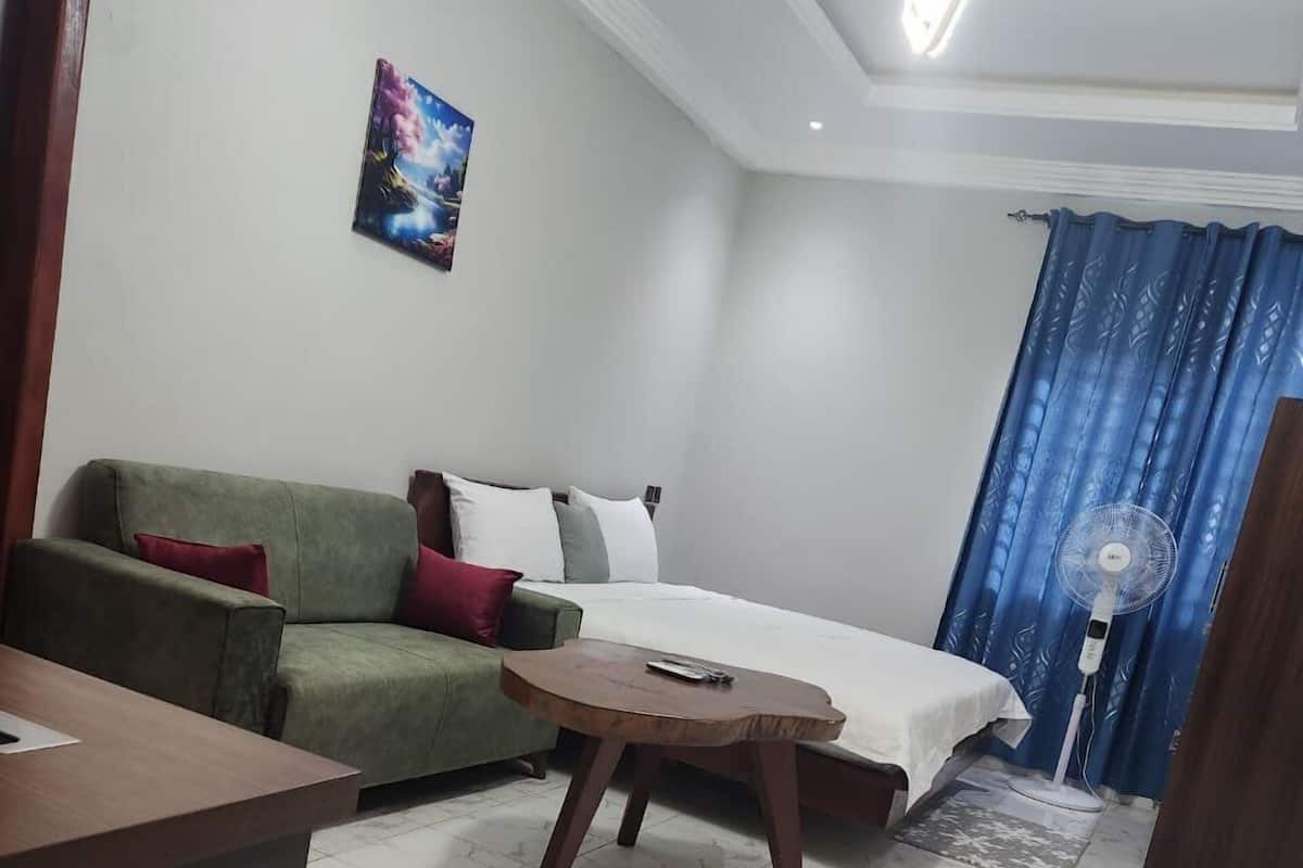 A small, well-lit room with a green sofa, a bed, a small round table, and a standing fan.