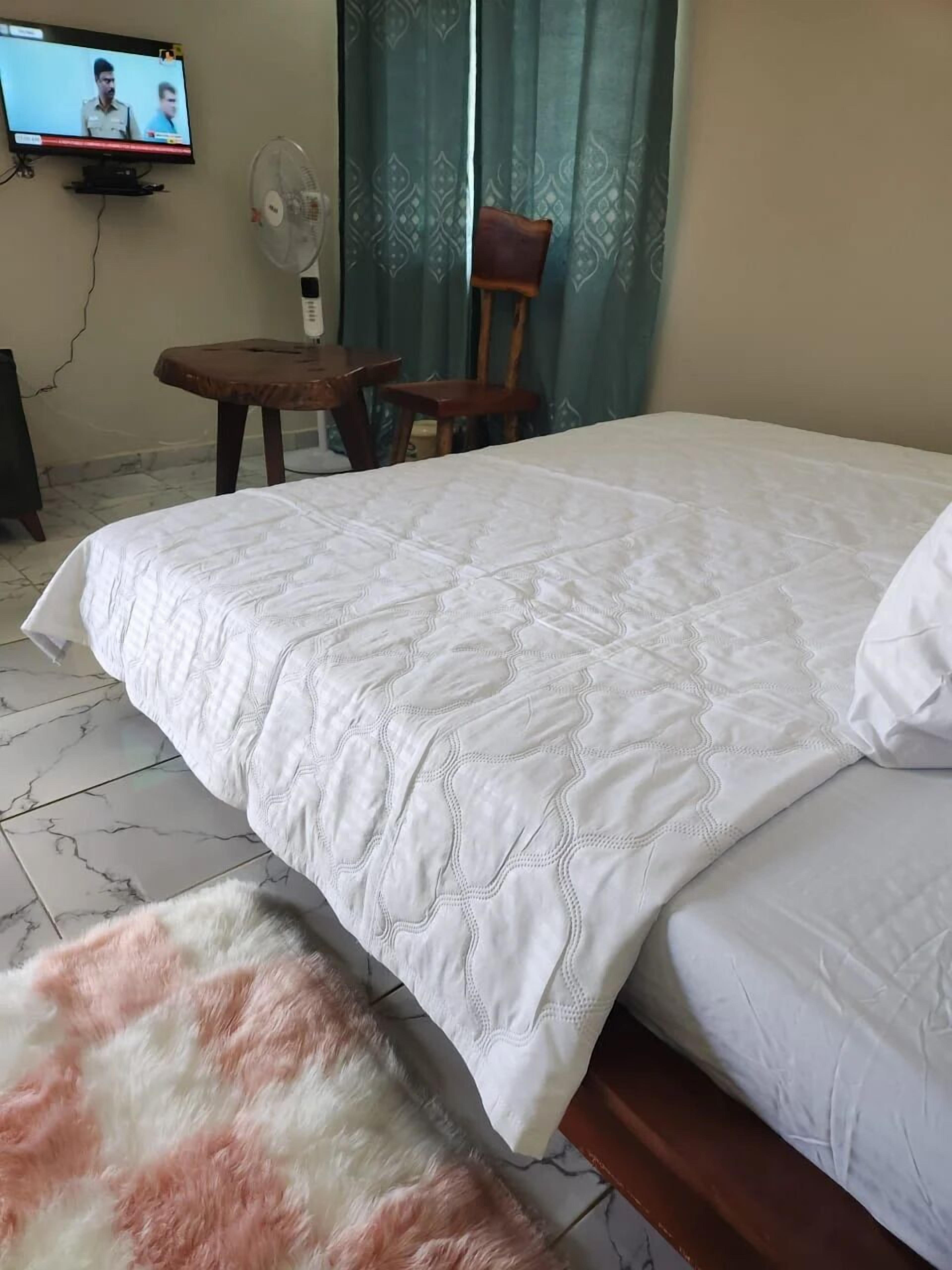 A hotel room with a large bed, a small table, a chair, a TV, and a wall-mounted fan.