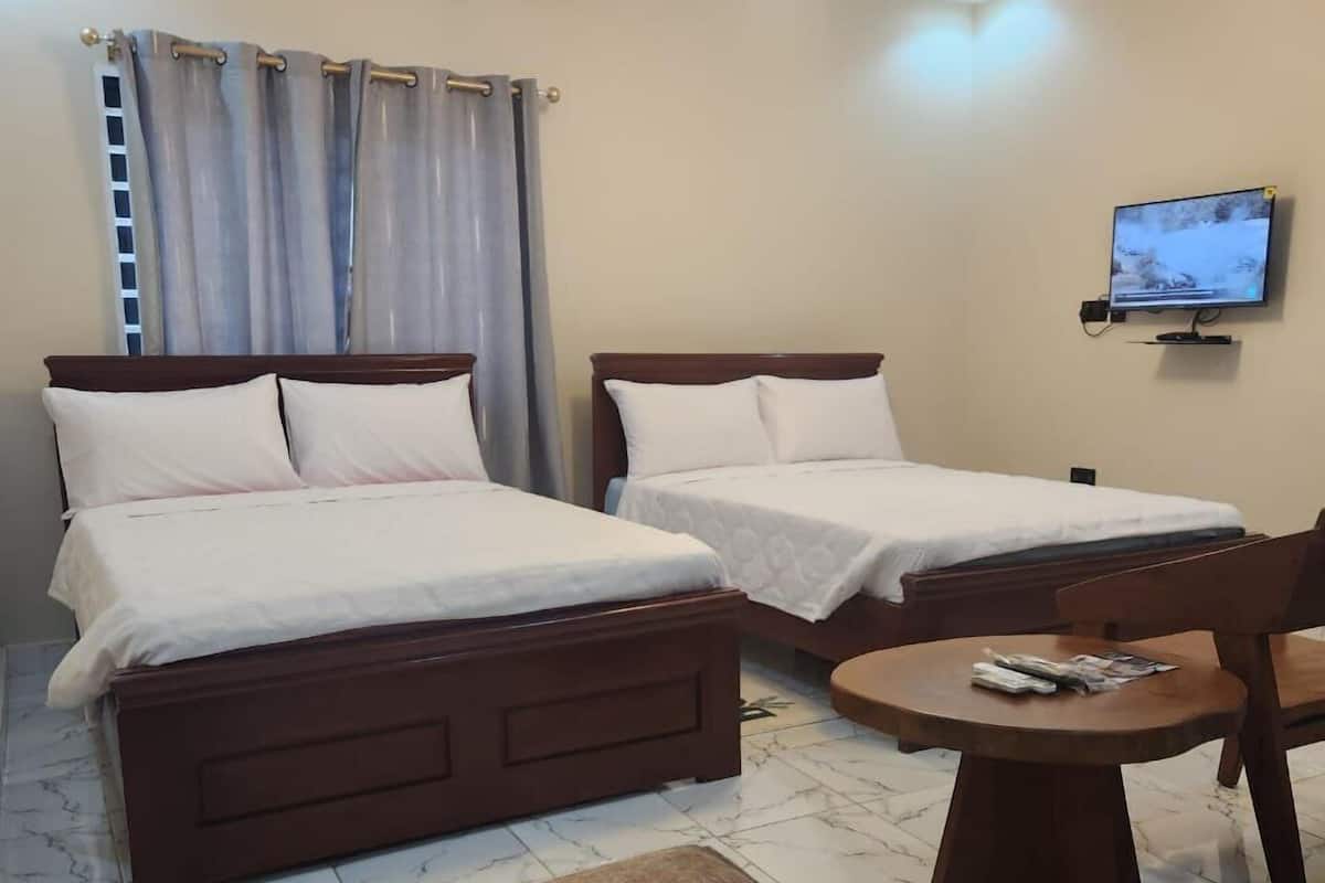A hotel room with two wooden beds, a small round table with a chair, a television on the wall, and a window with curtains.