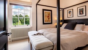 Ein Schlafzimmer mit großem Fenster, einem Bett mit Kopfteil, einem Nachttisch mit Lampe und gerahmten Bildern an der Wand.