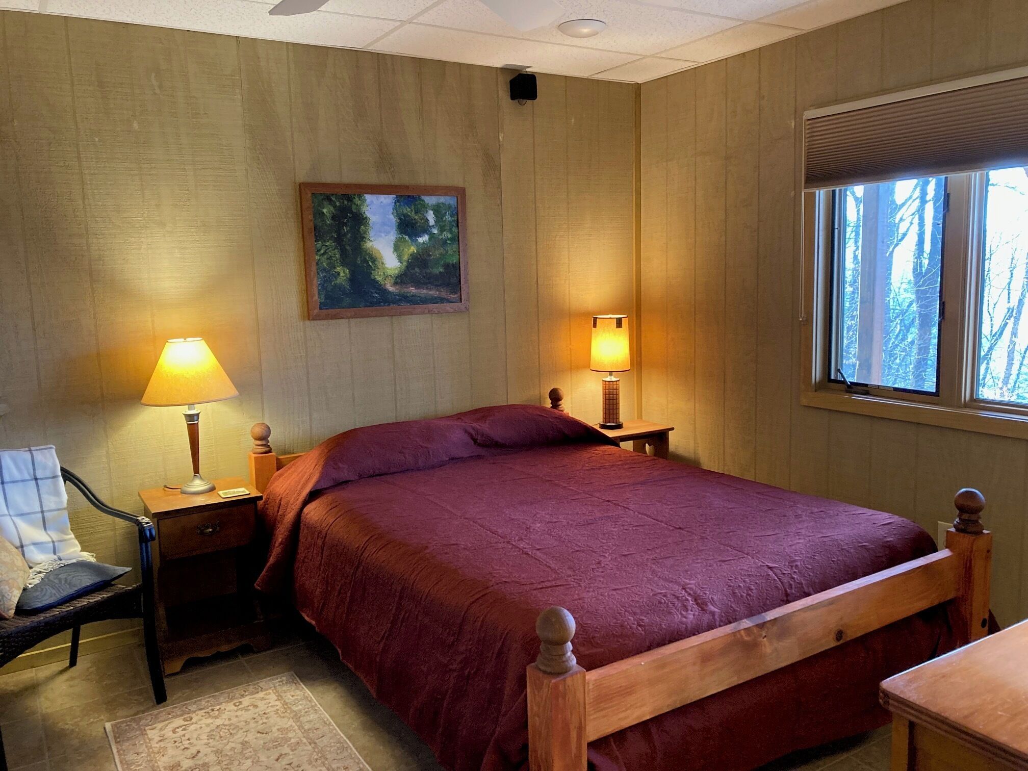 Un dormitorio con una cama de madera, dos lámparas de noche, una mesita de noche, una silla, una ventana con vistas a árboles y un cuadro en la pared.