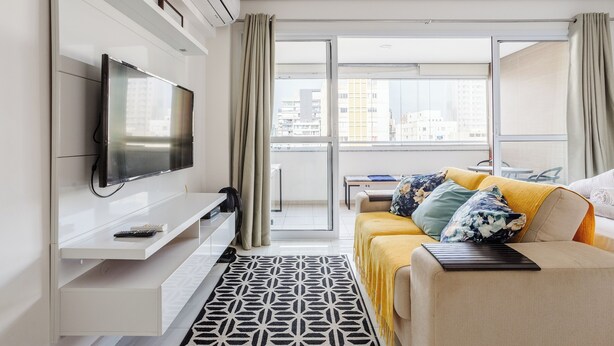 Een moderne hotelkamer met een grote flatscreen-tv aan een witte muur, een bank met geel en bloemrijk bedrukte kussens, een kamerbreed tapijt en een schuifdeur naar een balkon met uitzicht op de stad.