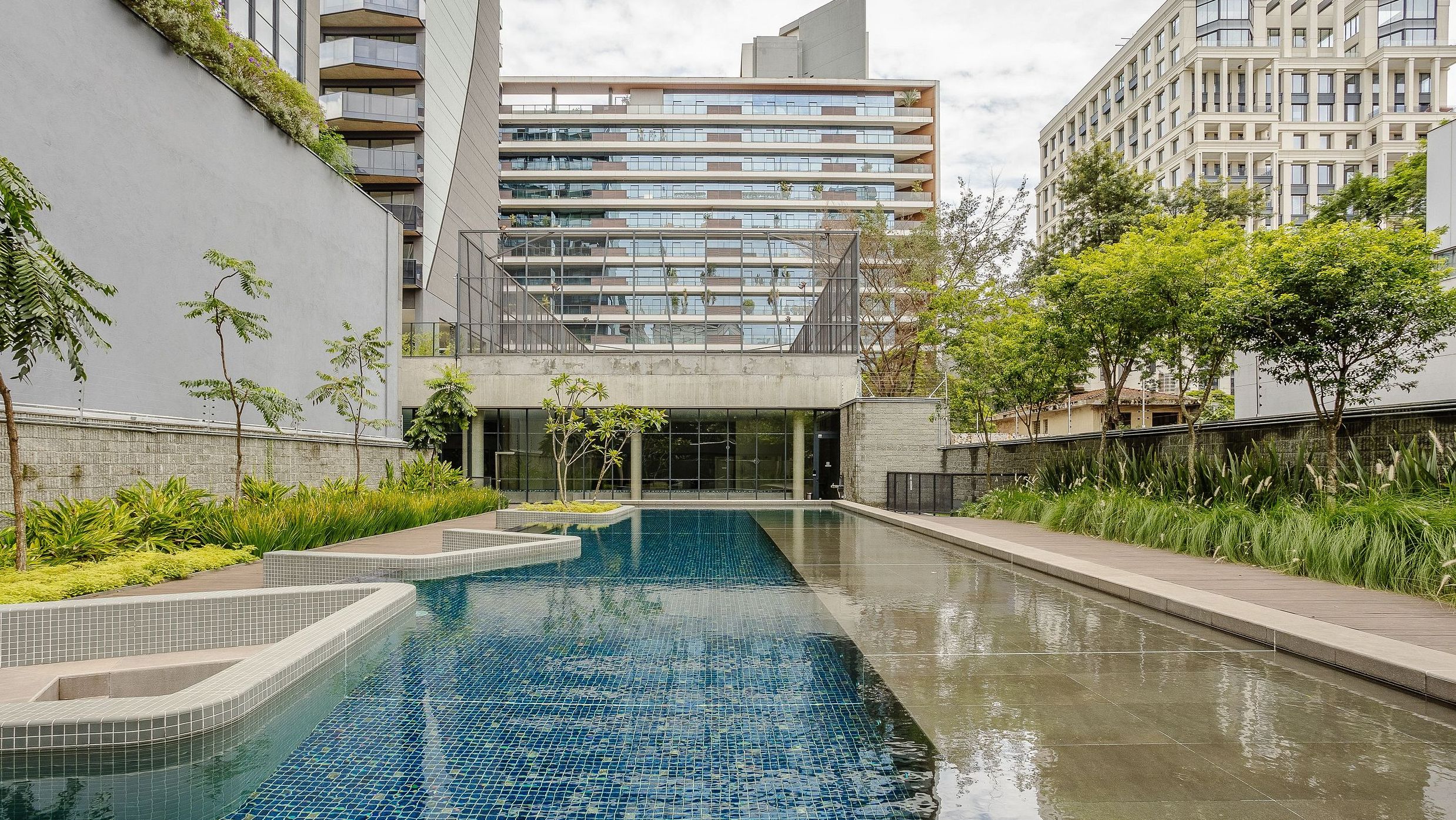 Un edificio moderno con piscina, rodeado de vegetación y otros edificios residenciales.