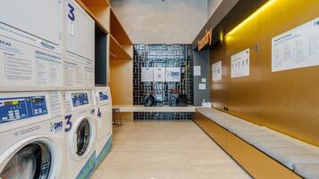 Une buanderie équipée de plusieurs machines à laver, un espace détente et des affiches d’information accrochées aux murs.