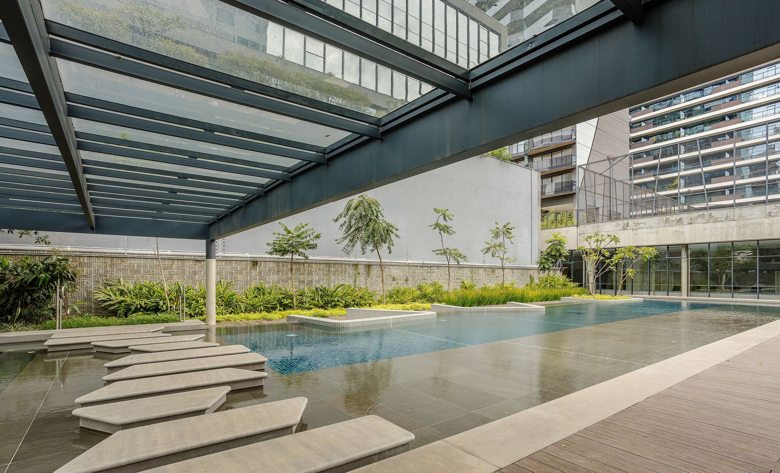A modern indoor pool area with a glass ceiling, surrounded by greenery and a view of a high-rise building.