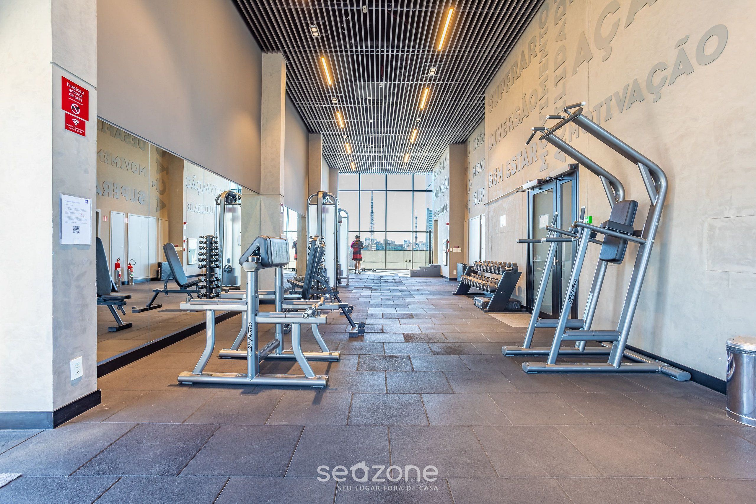Une salle de sport moderne dotée de divers appareils d’exercice, de grandes fenêtres et d’un plafond haut avec un éclairage linéaire.
