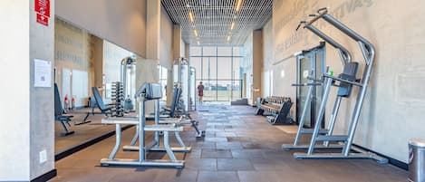 Un gimnasio moderno con diversos equipos de ejercicio, grandes ventanales y un techo alto con iluminación lineal.