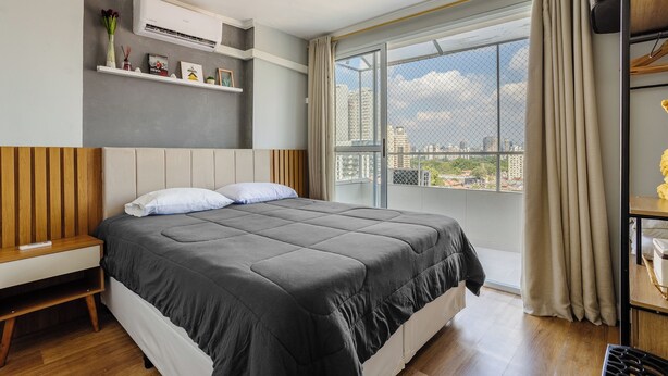 A modern bedroom with a large bed, a bedside table, a shelf with decorative items, and a view of the city through a large window. - Charming Apartment in Pinheiros - Cot0081 (São Paulo)